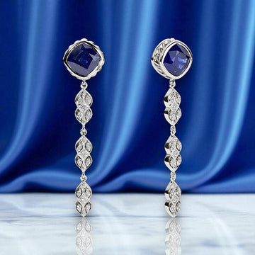 Pair of silver earrings with blue gemstones and diamond accents displayed on a blue satin fabric and marble surface background.