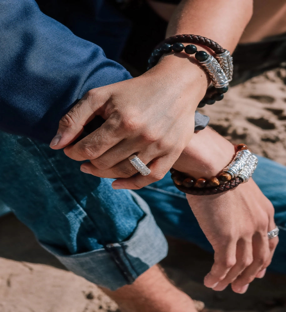 Close-up of a man’s hands wearing handcrafted silver and leather bracelets and a silver ring, sitting casually with rolled-up jeans on a sandy surface