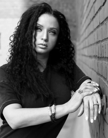 Black and white portrait of a woman wearing bracelets and rings, standing against a brick wall, showcasing the craftsmanship of handcrafted silver jewelry.