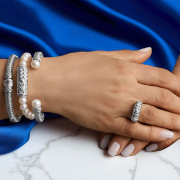 Studio Collection jewelry styled on model hand — sterling silver cuff, pearl bracelet with silver clasp, and engraved silver ring, displayed against blue satin and marble background.