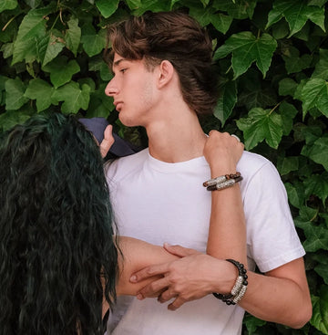 Young couple wearing handcrafted silver and leather bracelets, standing close in front of lush green ivy.