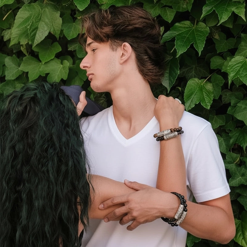 Lifestyle photo of a couple standing in front of lush green leaves, with both models wearing silver and beaded bracelets. The male model wears a fitted white V-neck T-shirt, while the female model’s arm wraps naturally around him, showcasing her jewelry. The image highlights the bracelets in a warm, intimate outdoor moment.