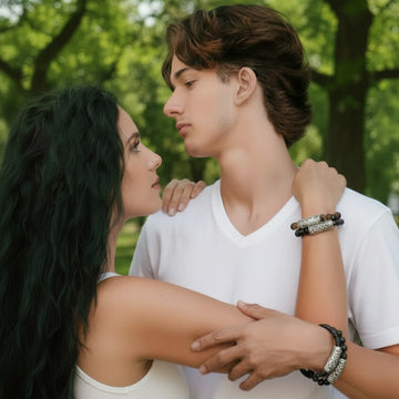 Couple standing closely together in a green park, both wearing Persian Shield silver bracelets paired with tiger-eye stones and black leather. The man wears two stacked bracelets while the woman’s hand rests on his shoulder, highlighting the engraved Persian Shield silver design.