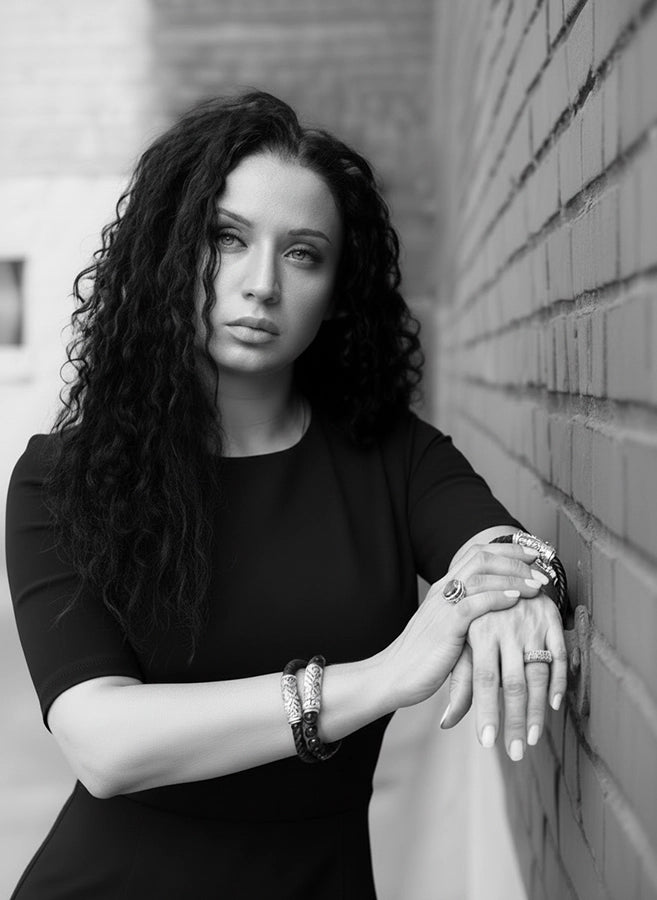 Black-and-white portrait of a woman in a fitted black dress leaning against a brick wall, with her hands crossed and showcasing multiple sterling-silver bracelets and rings. Her long curly hair frames her face as she looks slightly upward, creating a refined and elegant founder-style image.