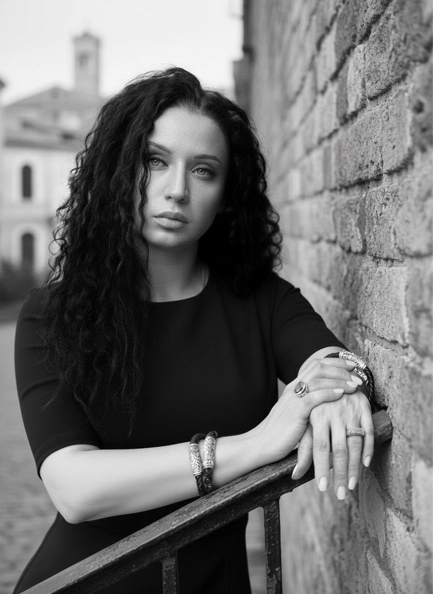 Black-and-white portrait of a woman in a fitted black dress leaning against a brick wall, with her hands crossed and showcasing multiple sterling-silver bracelets and rings. Her long curly hair frames her face as she looks slightly upward, creating a refined and elegant founder-style image.