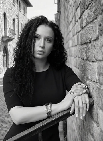 Black-and-white portrait of a woman in a fitted black dress leaning against a brick wall, with her hands crossed and showcasing multiple sterling-silver bracelets and rings. Her long curly hair frames her face as she looks slightly upward, creating a refined and elegant founder-style image.