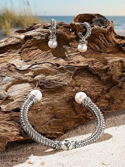 Sterling silver bracelet with Persian shield leaf–inspired metalwork and pearl ends, paired with matching pearl drop earrings, displayed on natural driftwood by the sea.