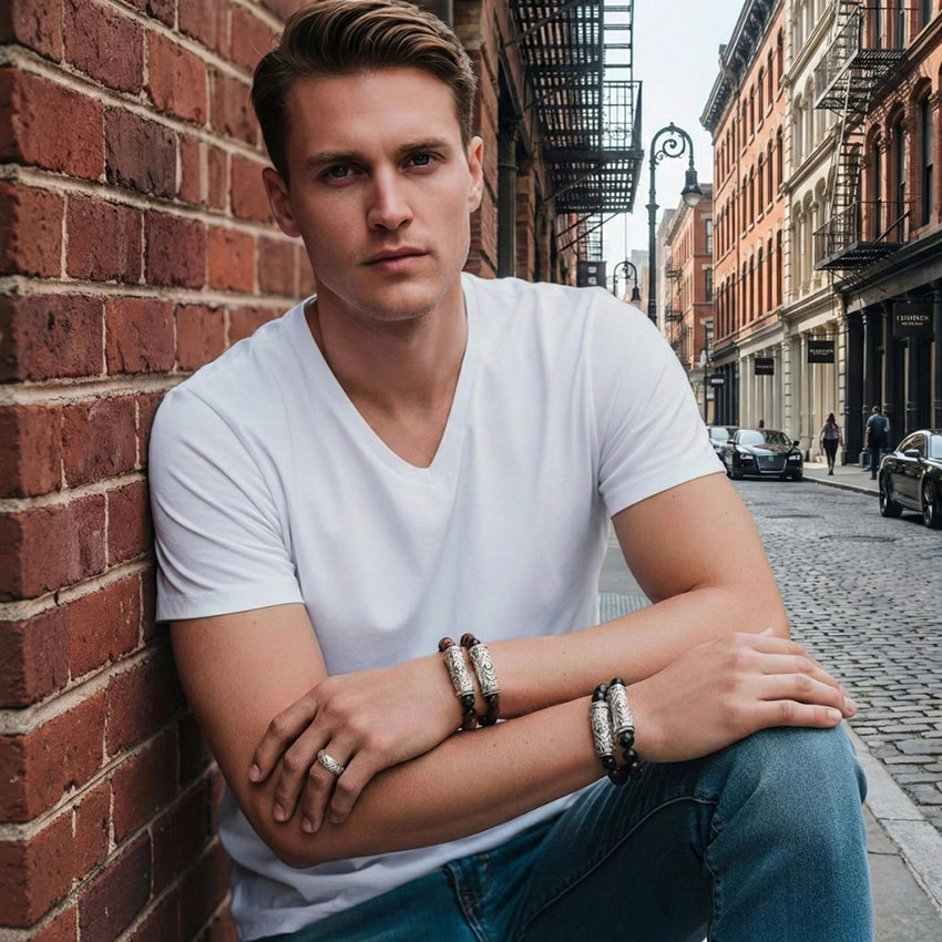 A man in a white V-neck T-shirt and jeans sitting against a brick wall in New York’s West Village, wearing Daneila Léon sculpted silver bracelets, beaded bracelets, and a silver ring, photographed in a crisp urban street setting