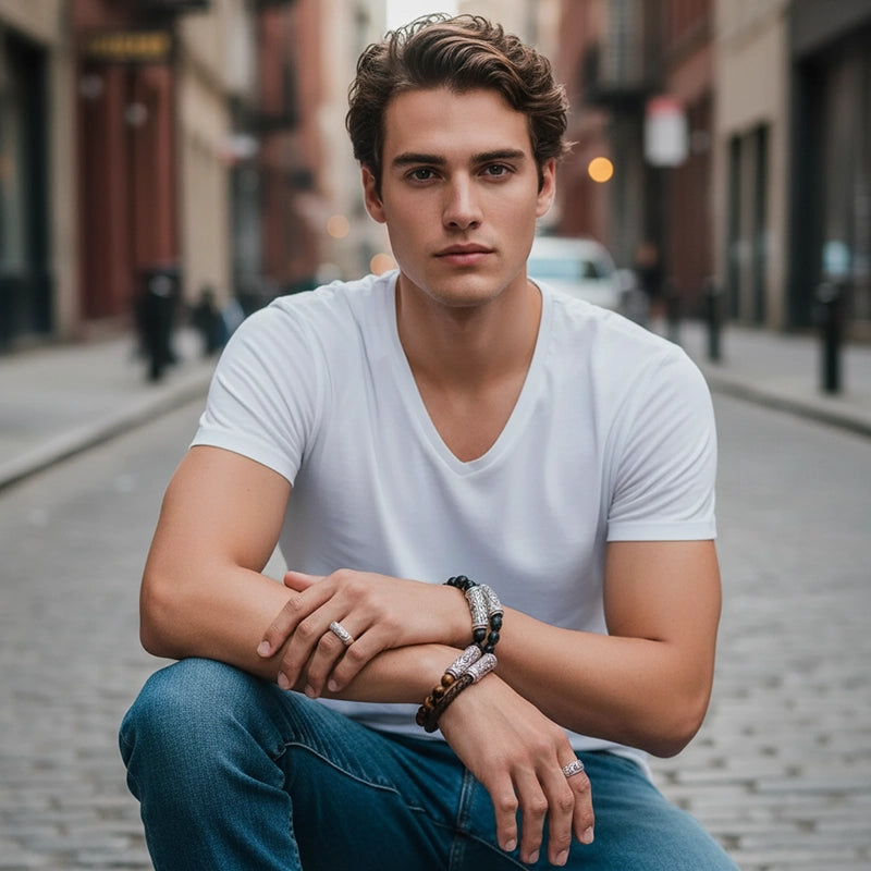 A man seated on a cobblestone NYC street wearing a white V-neck shirt and jeans, resting his arms on his knees while showcasing multiple handcrafted silver and beaded bracelets and a silver ring. The background features softly blurred urban buildings and warm street lighting.