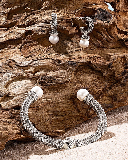 Pearl-accented silver cuff bracelet and matching hoop earrings displayed against textured driftwood and sand in a coastal-inspired jewelry still life.