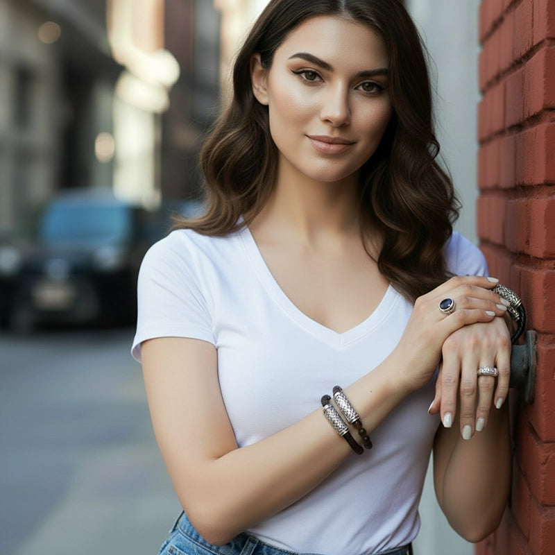A woman standing against a brick wall in a white V-neck top, softly smiling while displaying handcrafted silver and gemstone bracelets and rings. She poses with her hands gently crossed, showing detailed metalwork and polished dark beads. A softly blurred NYC street forms the background.
