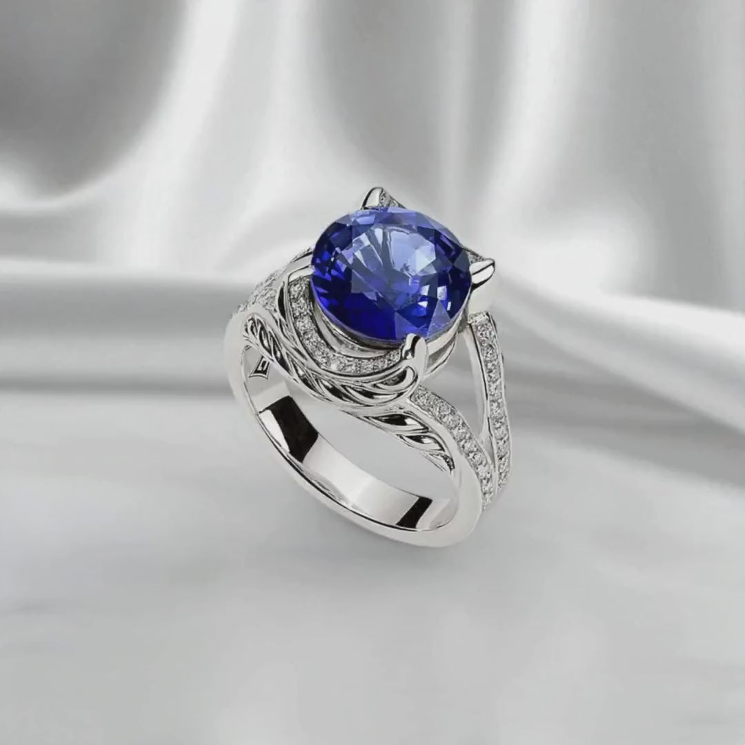 Silver ring with a vivid round blue sapphire center stone and diamond accents, shown on white satin fabric in a slow side-to-side loop. Soft diffused lighting highlights the handcrafted silver and gemstone design.