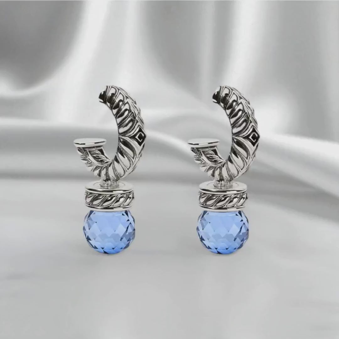Pair of silver earrings with round blue sapphire gemstones, shown on white satin fabric in a slow side-to-side loop. Soft diffused lighting highlights the sapphires’ deep blue color and the refined silver setting
