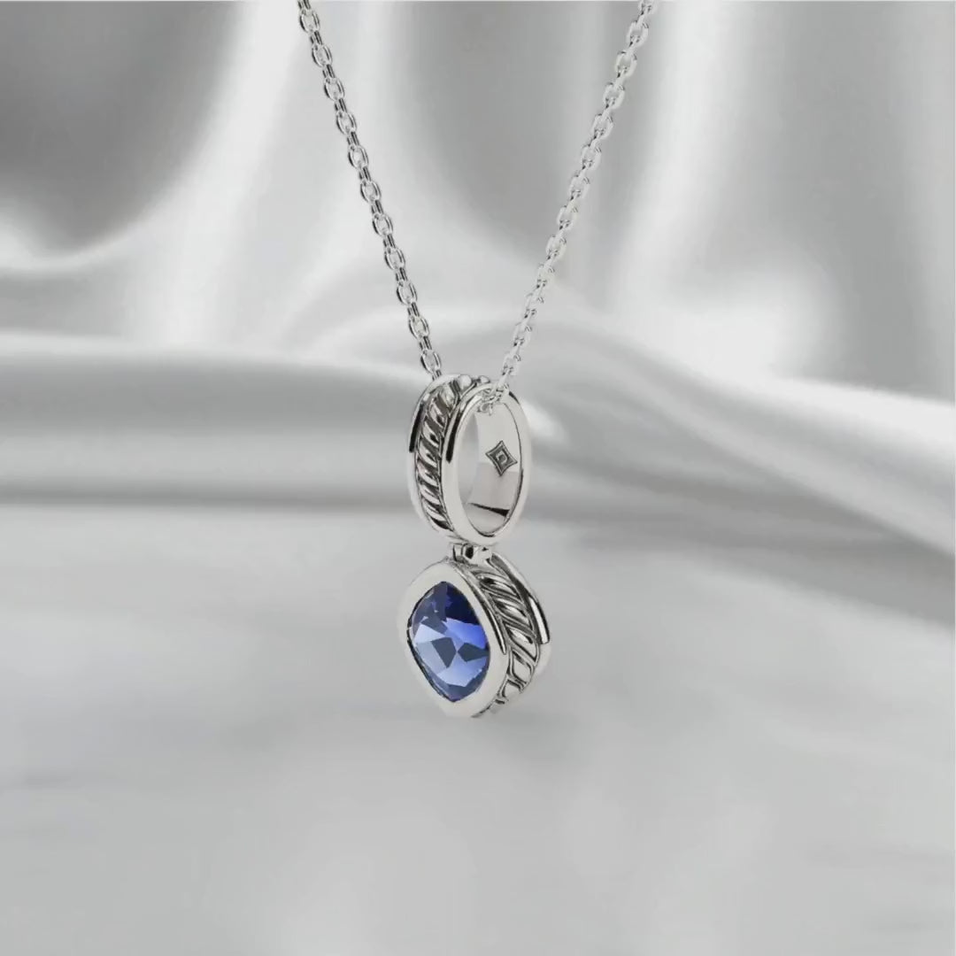 Sterling silver pendant necklace with lab-grown blue sapphire drop, displayed on a white satin backdrop with soft studio lighting and gentle movement to reveal its polished finish