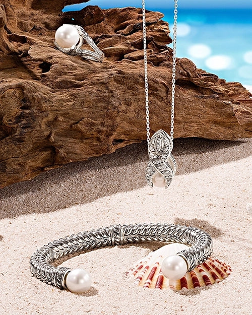 Silver pearl cuff bracelet, pendant necklace, and statement ring displayed on sand and driftwood with a soft ocean backdrop.